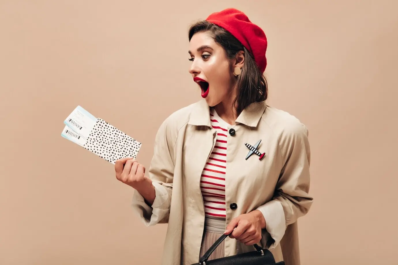 Удивленная женщина в красном берете и бежевом жакете смотрит на билеты. Стильная леди с яркими губами в берете, бежевом плаще и полосатом свитере позирует.