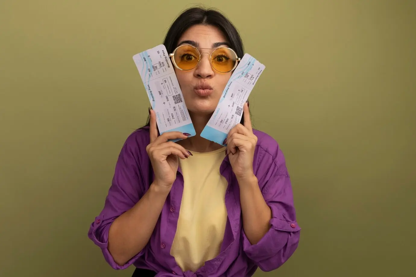 Впечатлённая симпатичная брюнетка в солнцезащитных очках держит авиабилеты близко к лицу на фоне оливково‑зелёной стены.