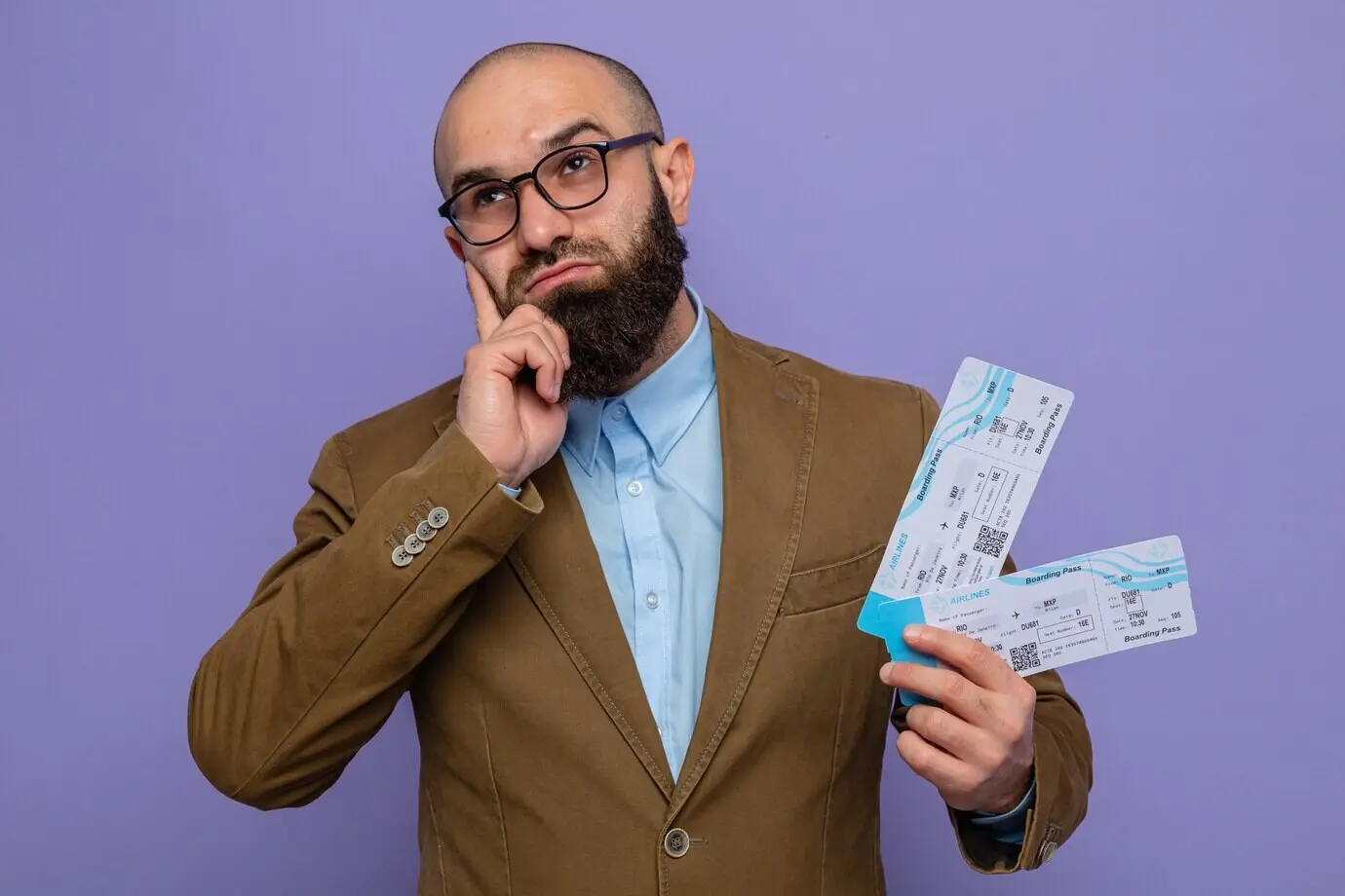 Бородатый мужчина в коричневом костюме и очках, держащий в руках авиабилеты, с недоумением смотрит вверх, стоя на фиолетовом фоне.