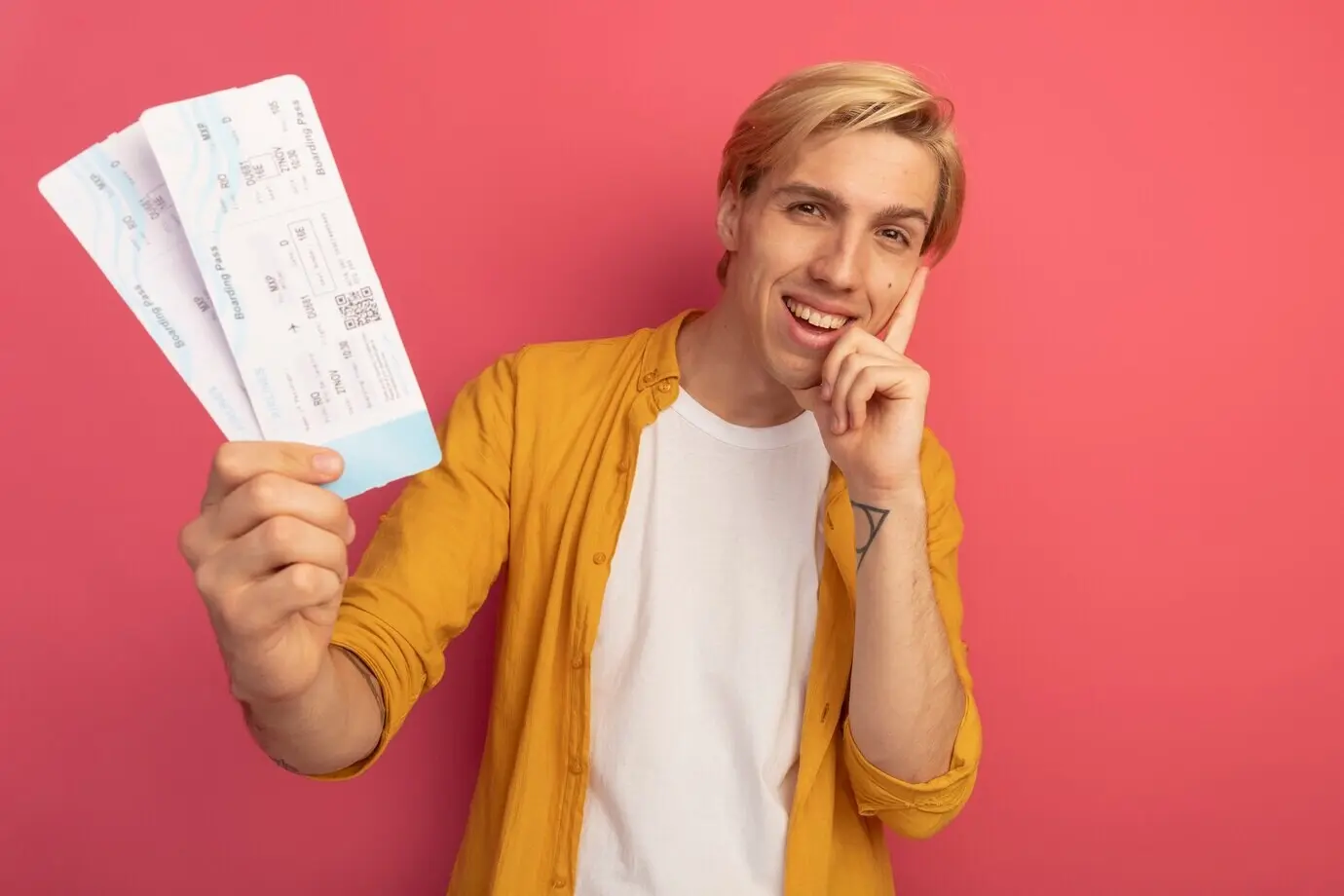 Улыбающийся молодой блондин в жёлтой футболке, держащий билеты и прикладывающий руку к щеке, изолированный на розовом фоне с местом для текста.